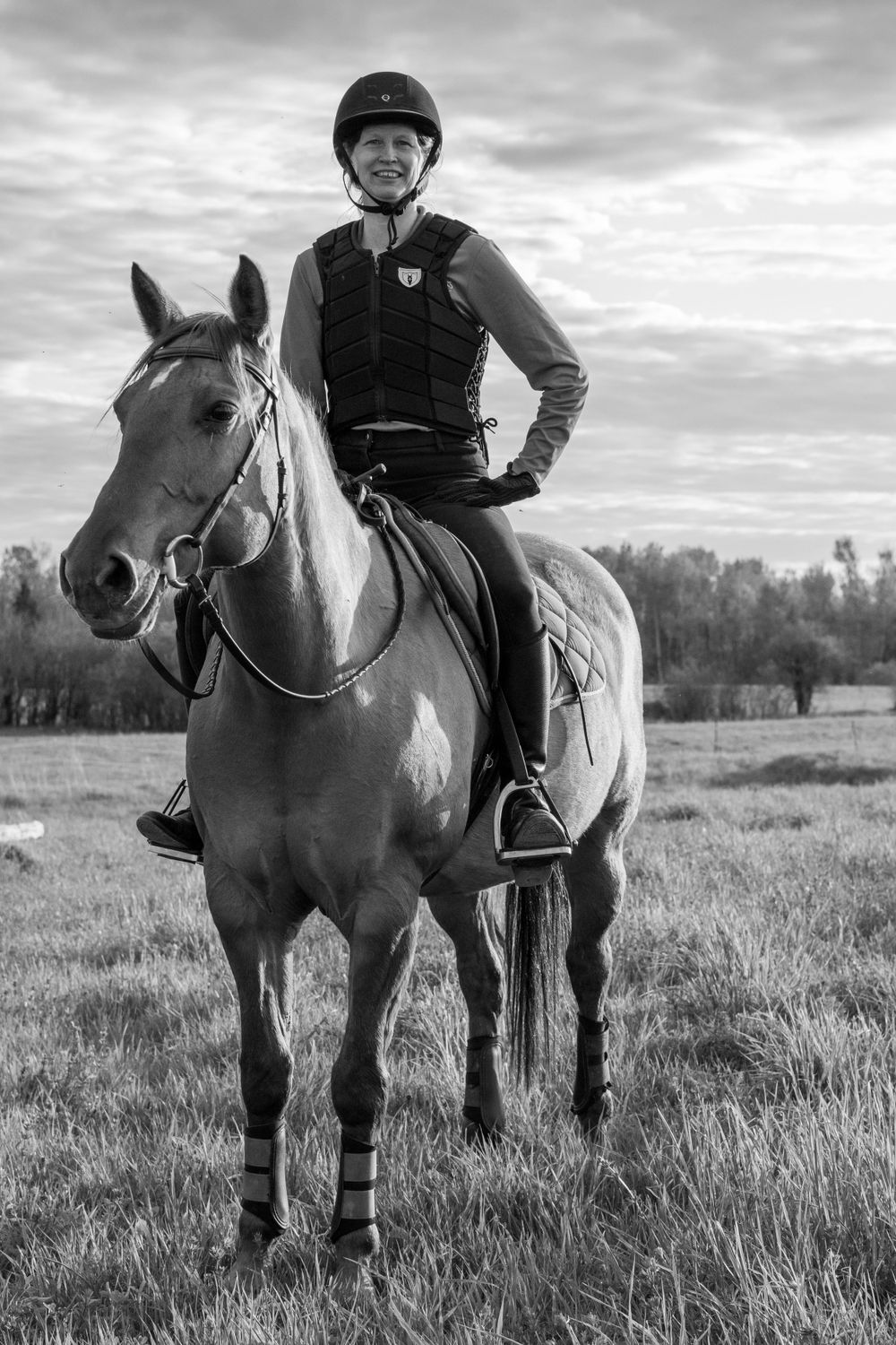 A black and white photo shows a rider on horseback in an open field with cloudy skies above.