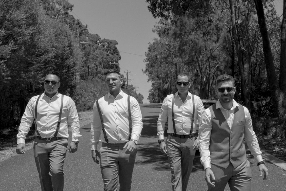 Four people in suspenders and formal wear walking down a tree-lined path.