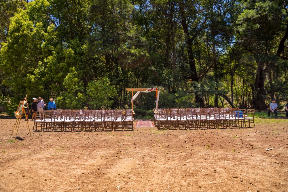Outdoor wedding ceremony setup with wooden arch and white chairs arranged in rows among tall trees.