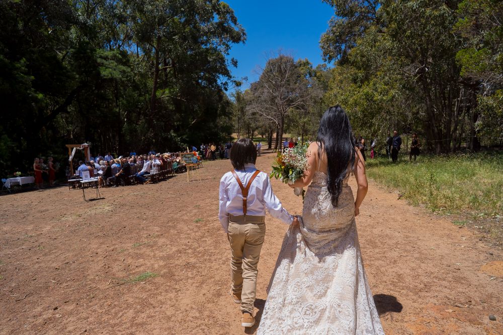 Wedding couple walking down dirt path with trees and guests in background.