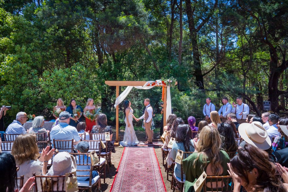 Wedding ceremony in progress under wooden arch with red carpet aisle and guests seated outdoors.