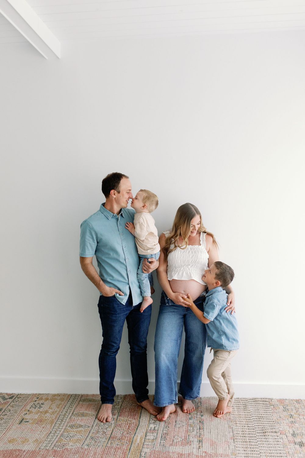A maternity family photo against a white wall with everyone wearing casual clothing and standing barefoot on a vintage rug.