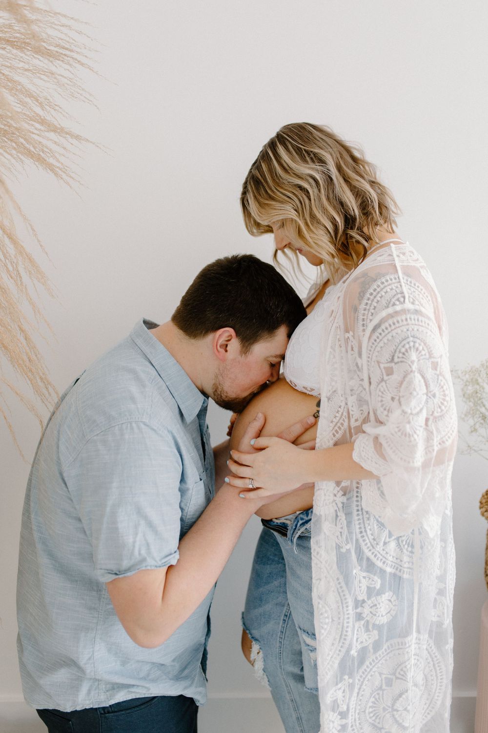 An intimate maternity moment captured against a white wall with decorative pampas grass.