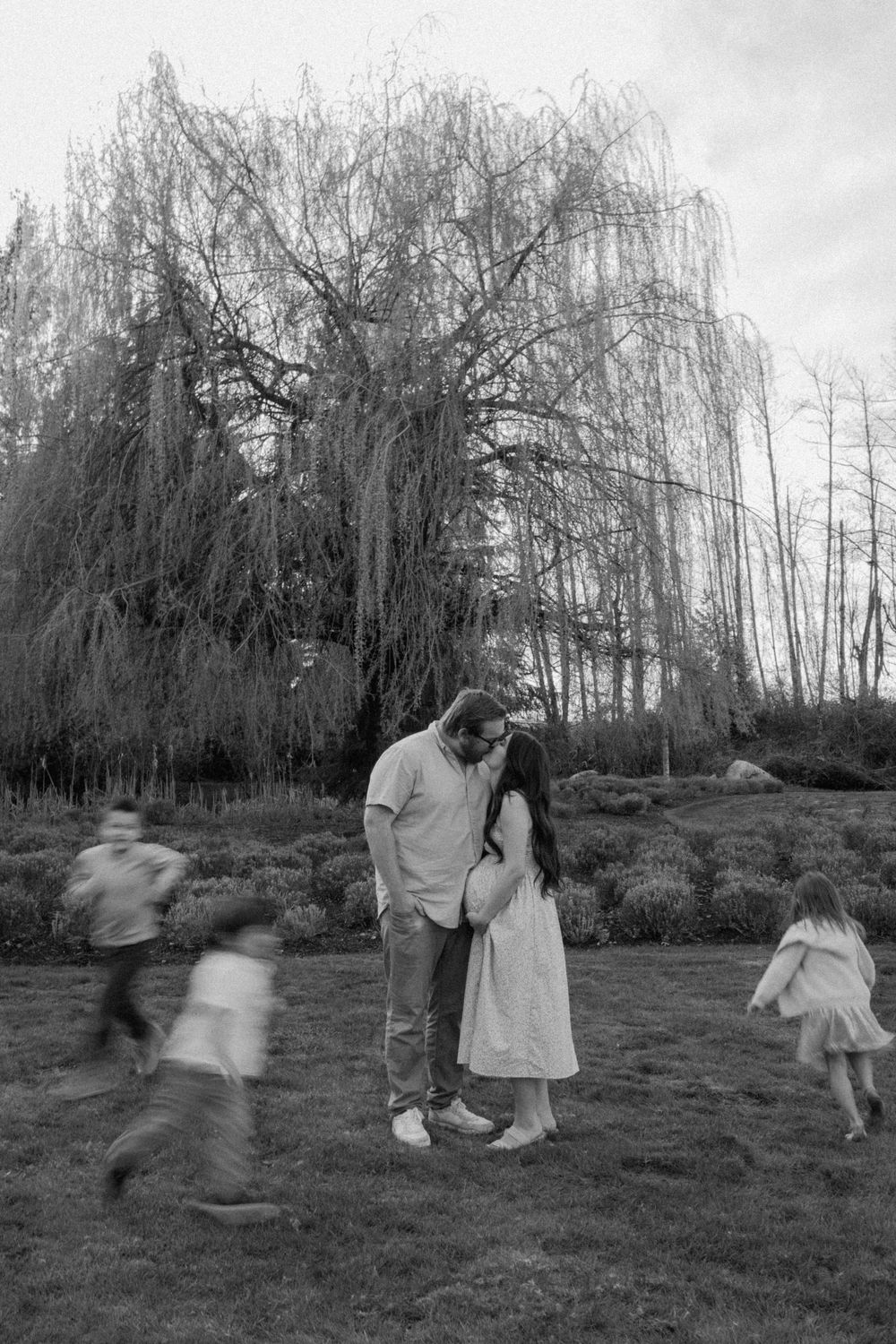 Black and white photo of couple embracing under weeping willow tree with motion-blurred children.