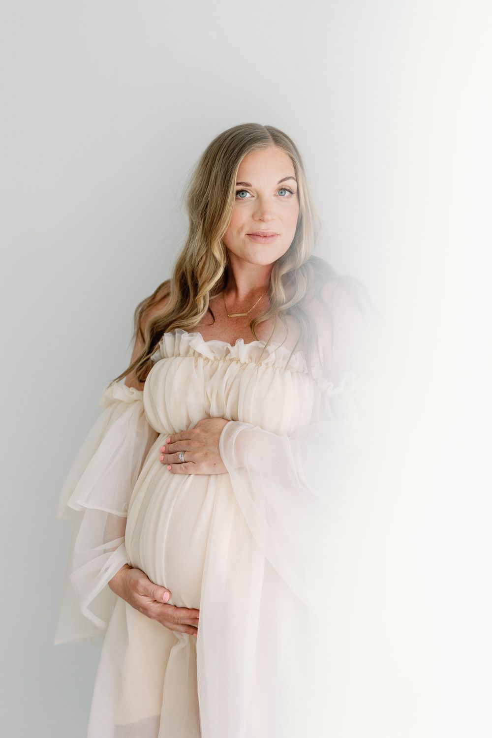 Ethereal maternity portrait with woman in flowing white dress against light gray background.
