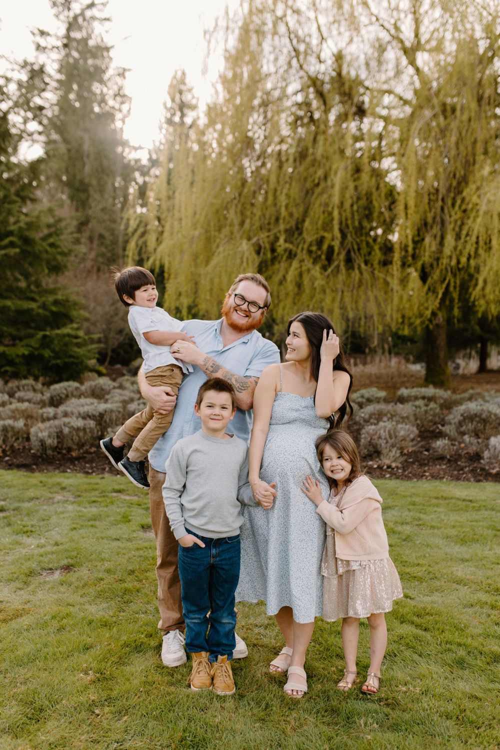 Family outdoors posing together in front of large weeping willow trees at sunset.