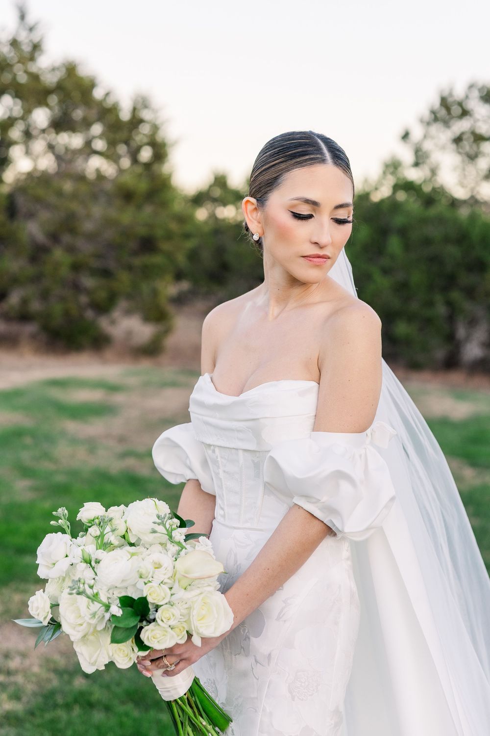 Bride in elegant off-shoulder wedding dress with puff sleeves holds white floral bouquet during outdoor sunset photo session.