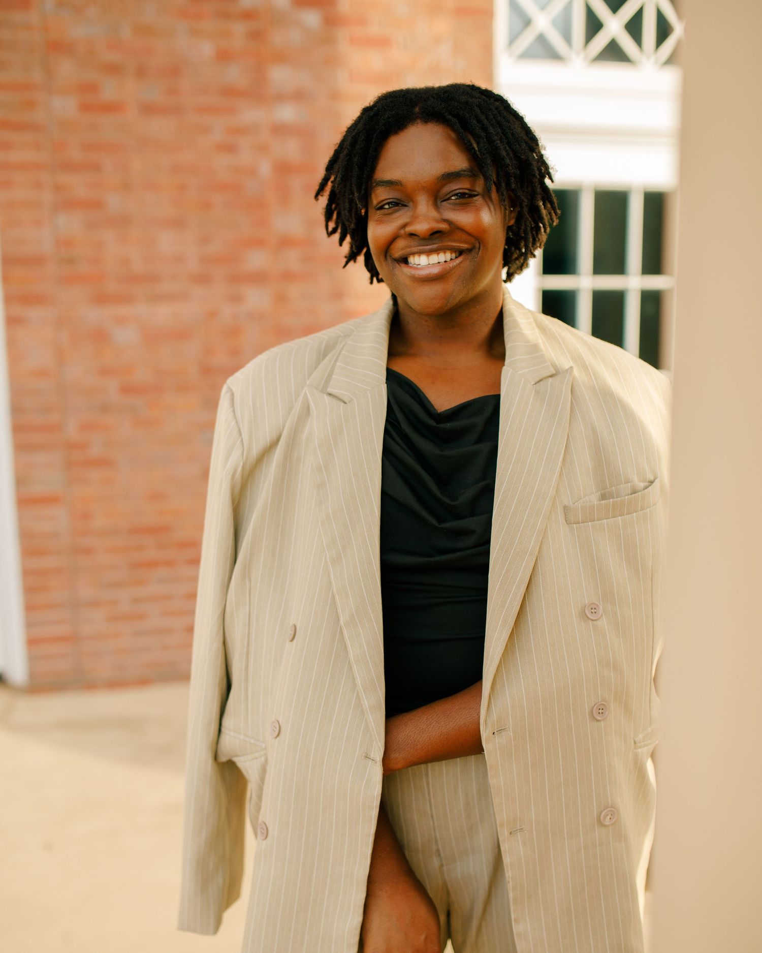 Person wearing an oversized beige blazer smiles warmly against a brick building backdrop.