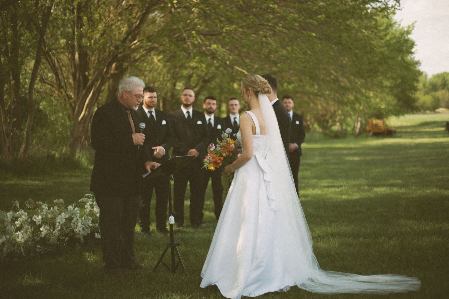 An outdoor wedding ceremony with a bride in white dress and officiant surrounded by trees and flowers.