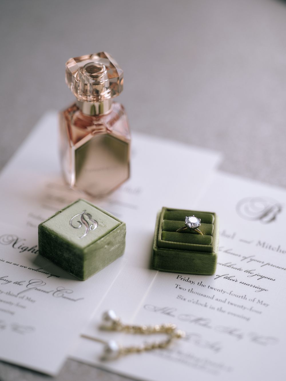 Wedding details with perfume bottle, green ring box, and invitation on white surface. Photoshoot by Venice Photographer Alina Indi