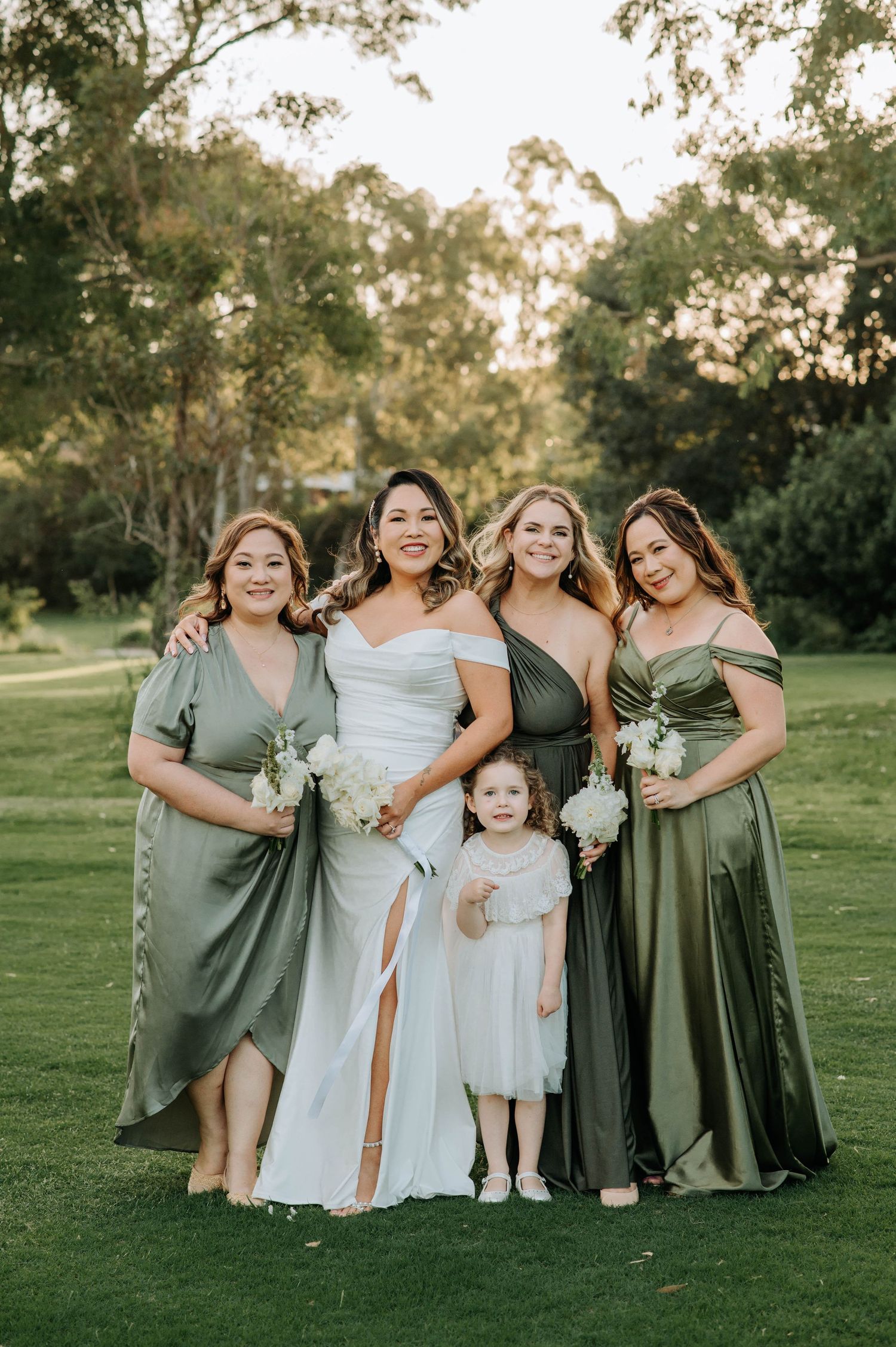 Bridal party in sage and olive green dresses pose together on a lush garden lawn.
