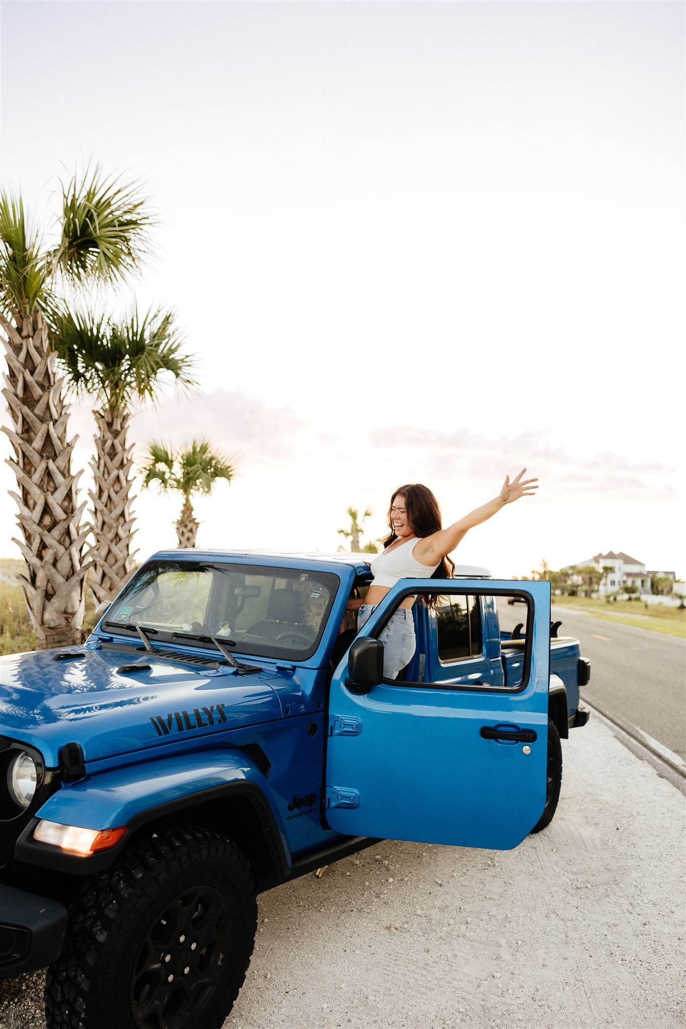 Woman in white top celebrating in bright blue Jeep Wrangler with palm trees.