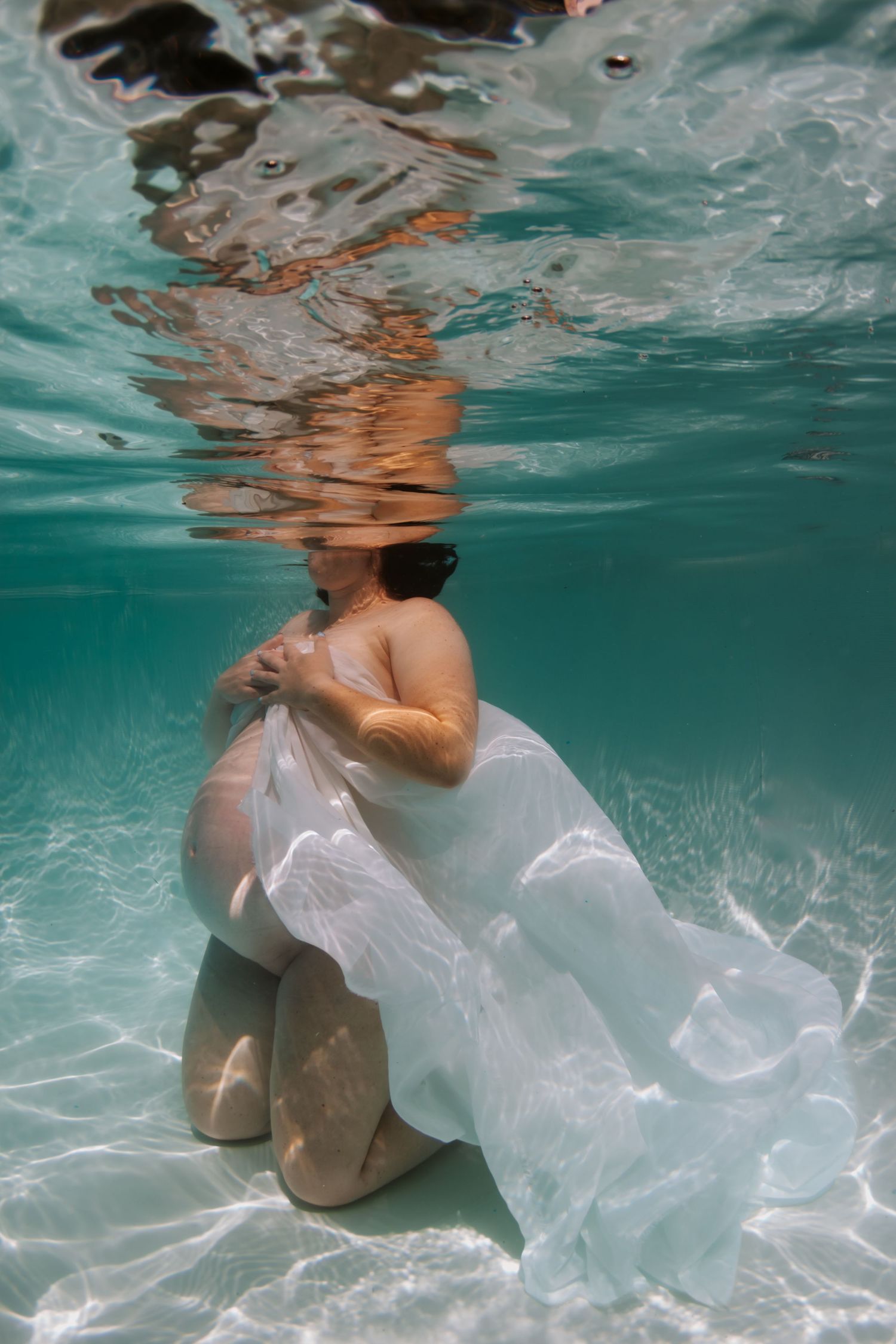 Pregnant woman kneeling underwater holding white fabric, serene maternity photo in clear turquoise water.