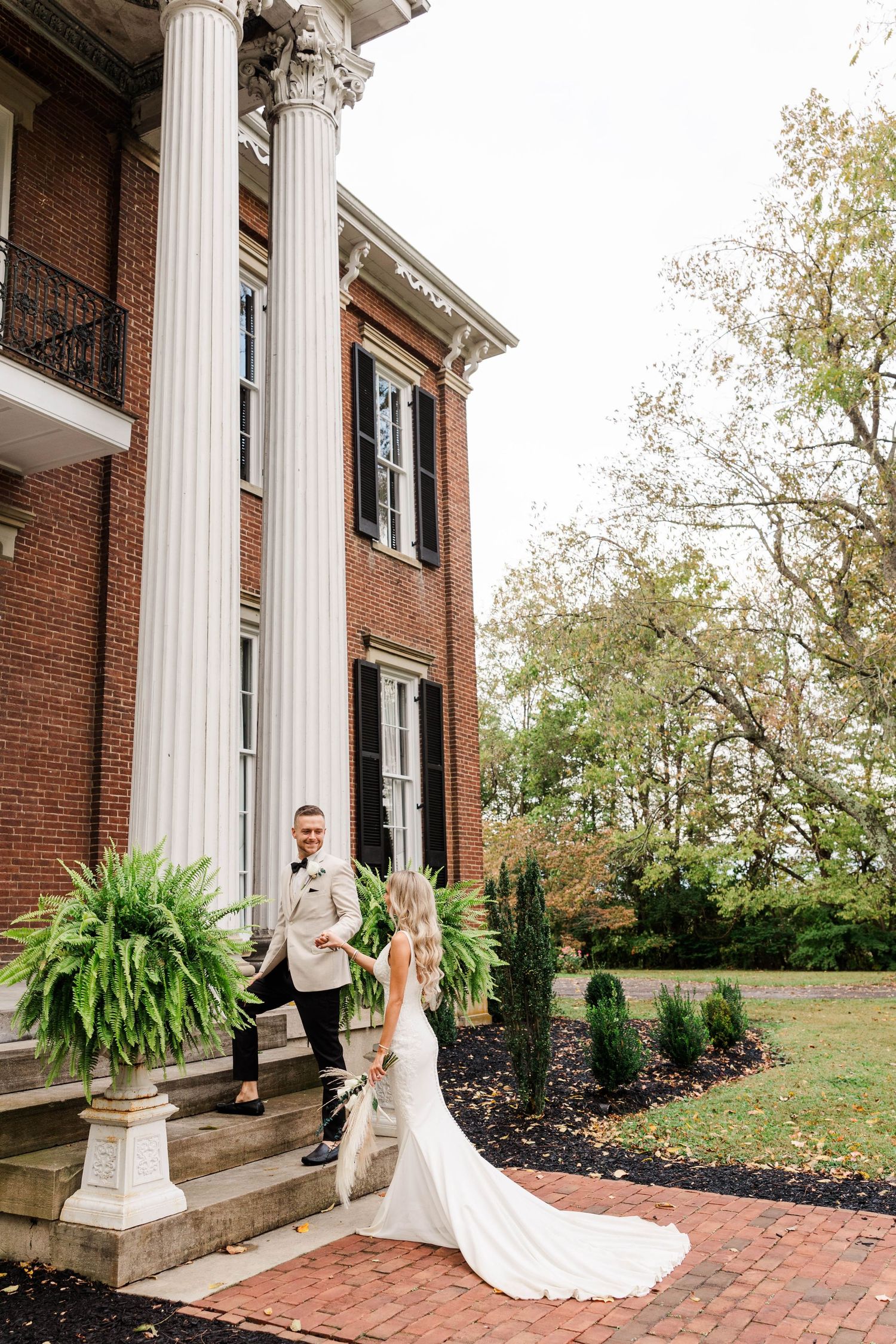 Elegant wedding couple on brick steps of historic mansion with white columns.