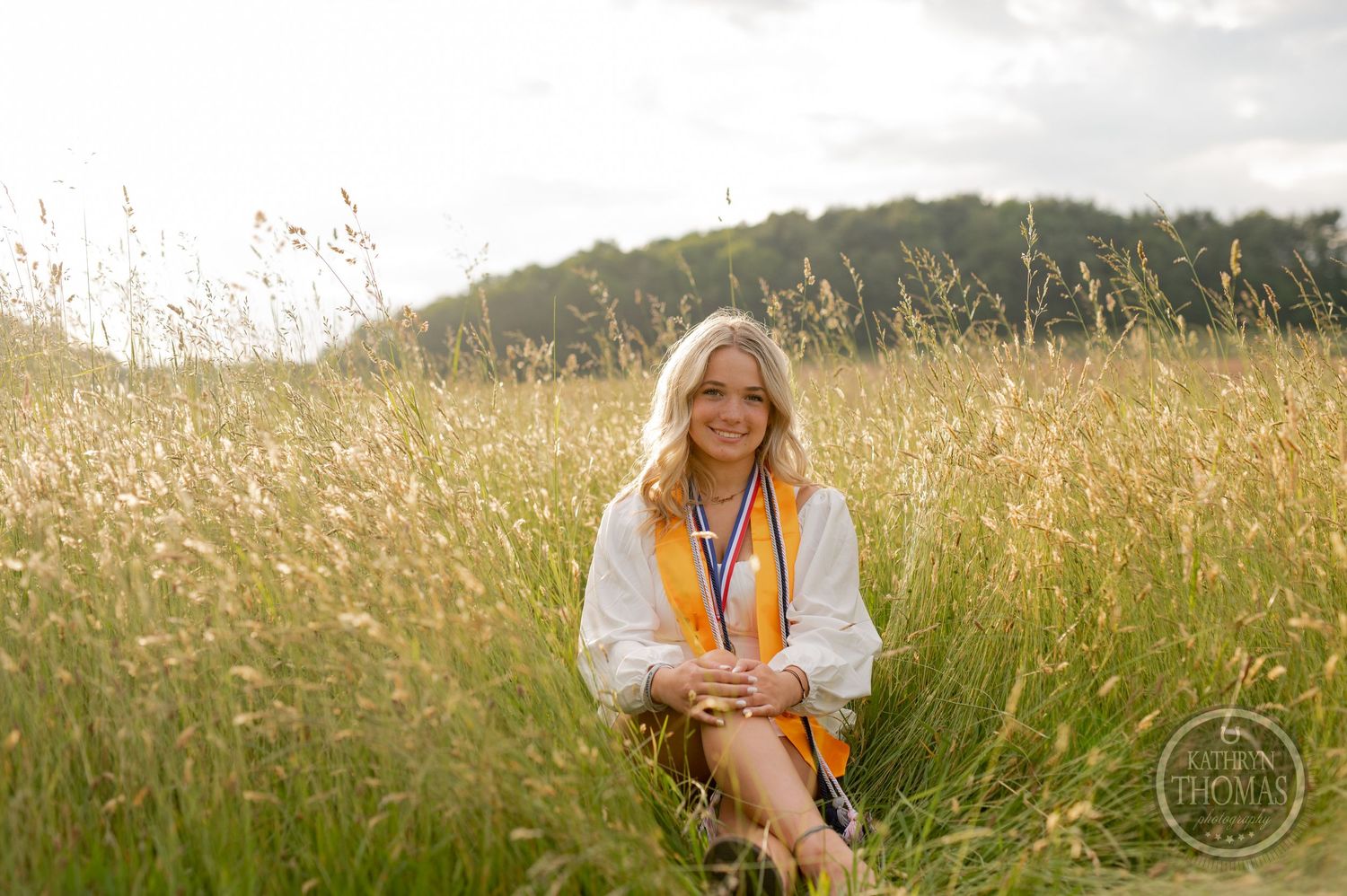 Graduate poses in a golden wheat field wearing a white top and academic regalia during sunset photo session.