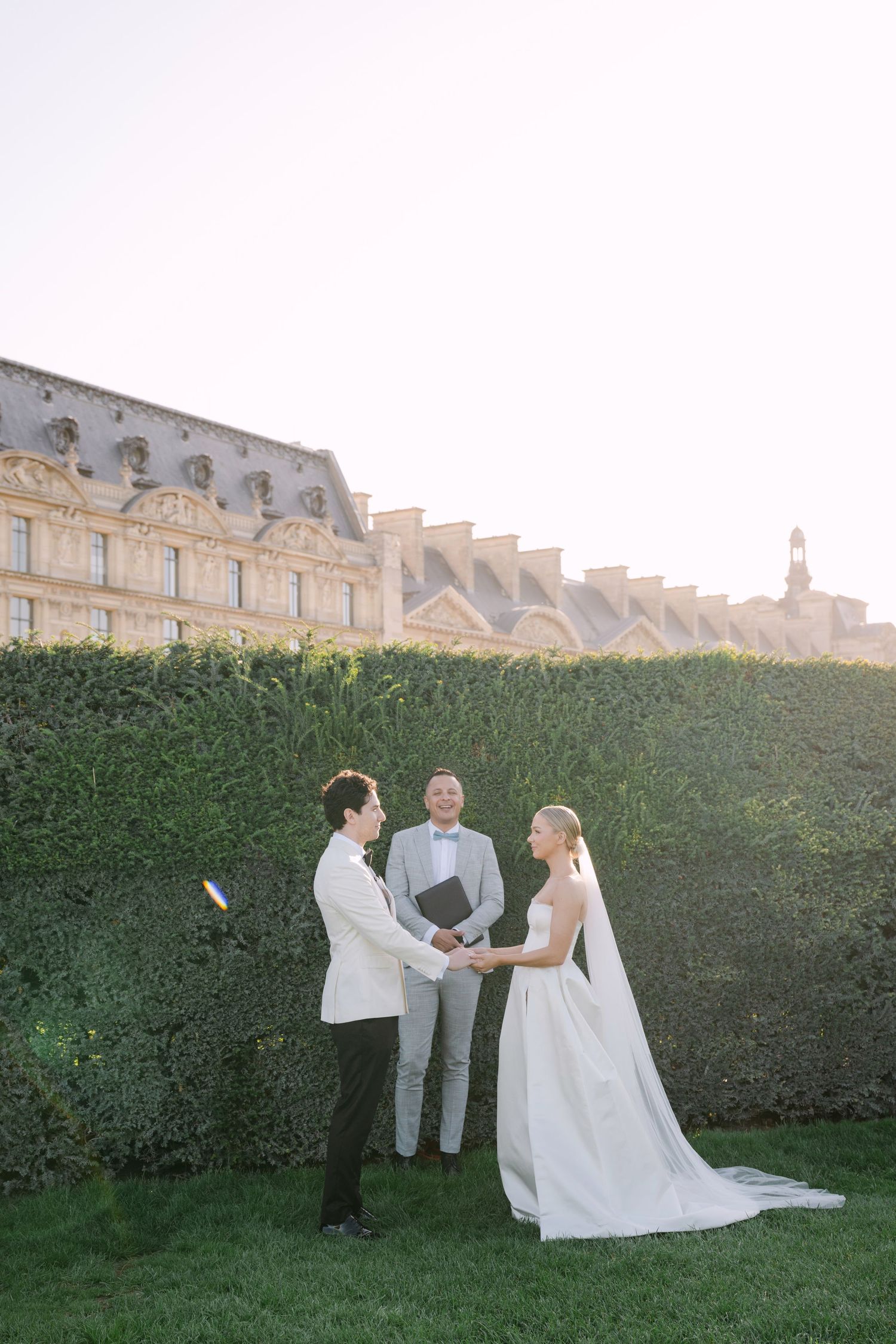 Couple in formal attire poses in front of the iconic Louvre Museum in Paris on a sunny day.