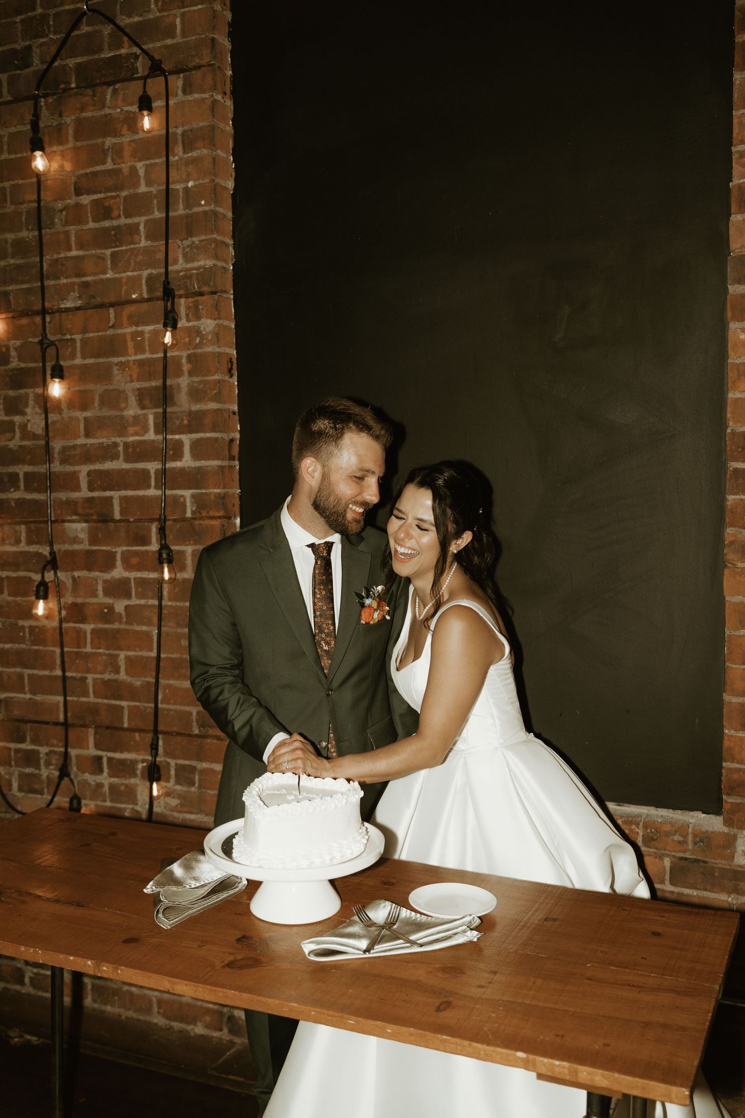 A couple in wedding attire cuts their white wedding cake together at a rustic venue with exposed brick walls and hanging lights.