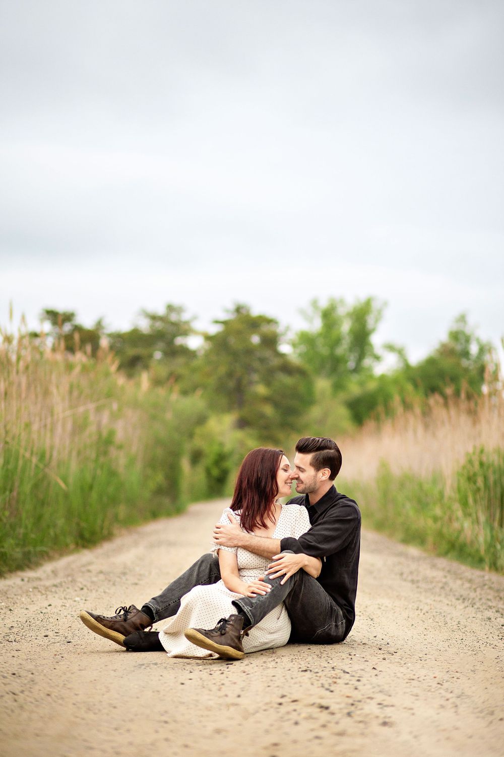 Two people sit together intimately on a dirt path surrounded by tall grass and trees on an overcast day.