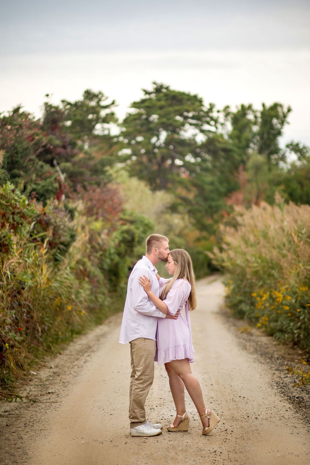 A romantic couple embracing on a dirt path surrounded by lush greenery and wildflowers.
