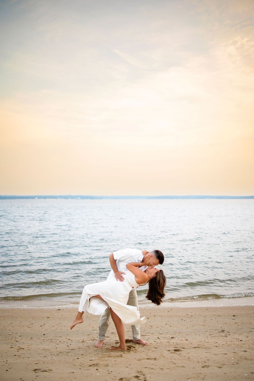 Two people share a romantic embrace and dance together on a peaceful beach at sunset with gentle waves in the background.