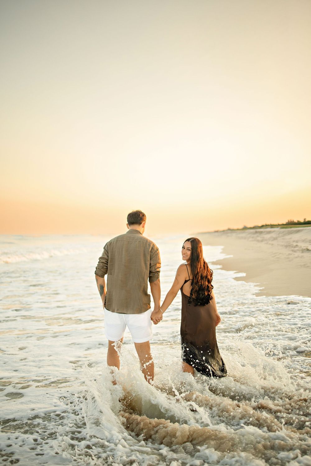 Couple holding hands walking along a sandy beach shore at golden sunset, waves splashing around their feet.