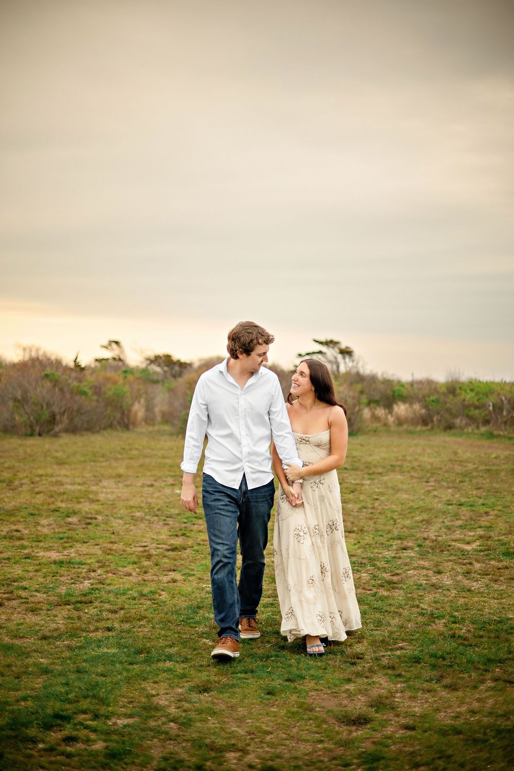A couple walks hand-in-hand through a grassy field, smiling at each other in casual elegant attire at sunset.