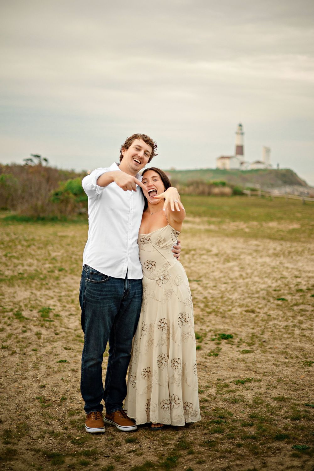 Happy couple laughing and pointing on a sandy beach with a lighthouse in the background on a cloudy day.