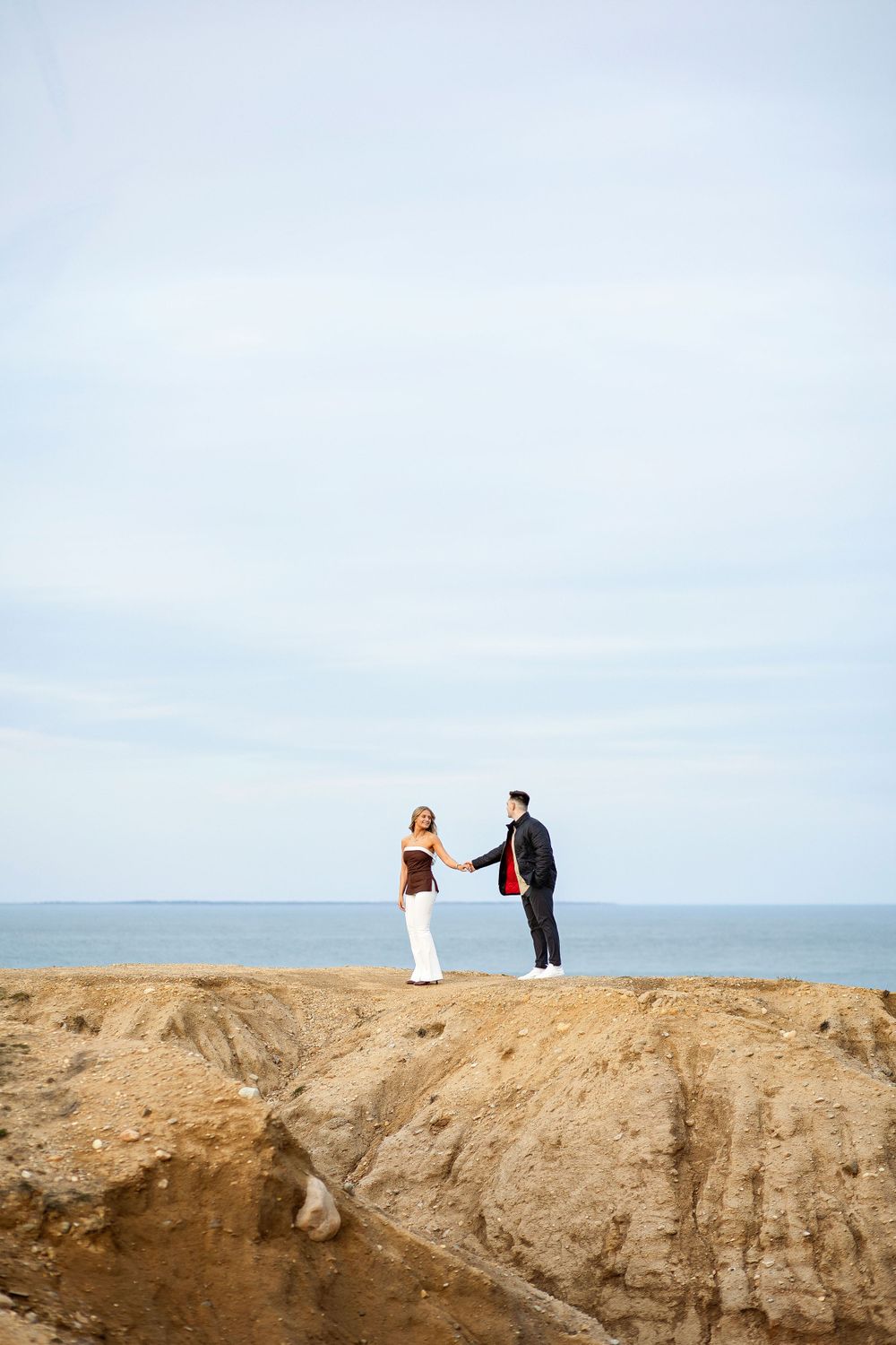 Two people stand together holding hands at the edge of a coastal cliff overlooking the ocean on a bright day.