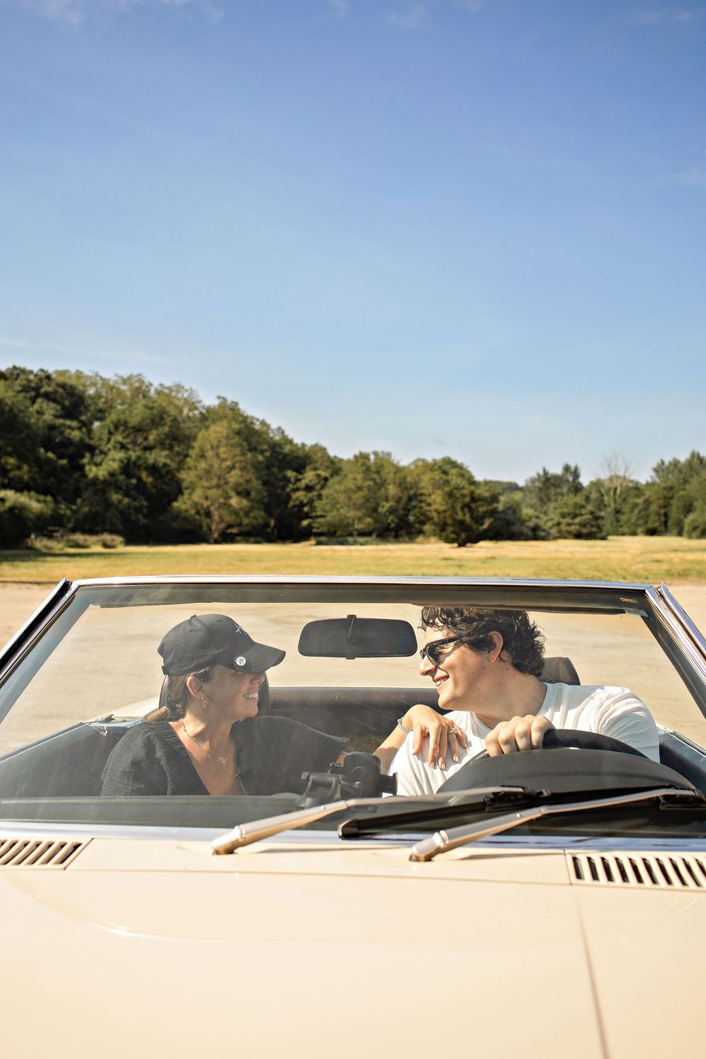 A couple enjoying a sunny road trip in a vintage cream convertible car, surrounded by green trees and blue sky.