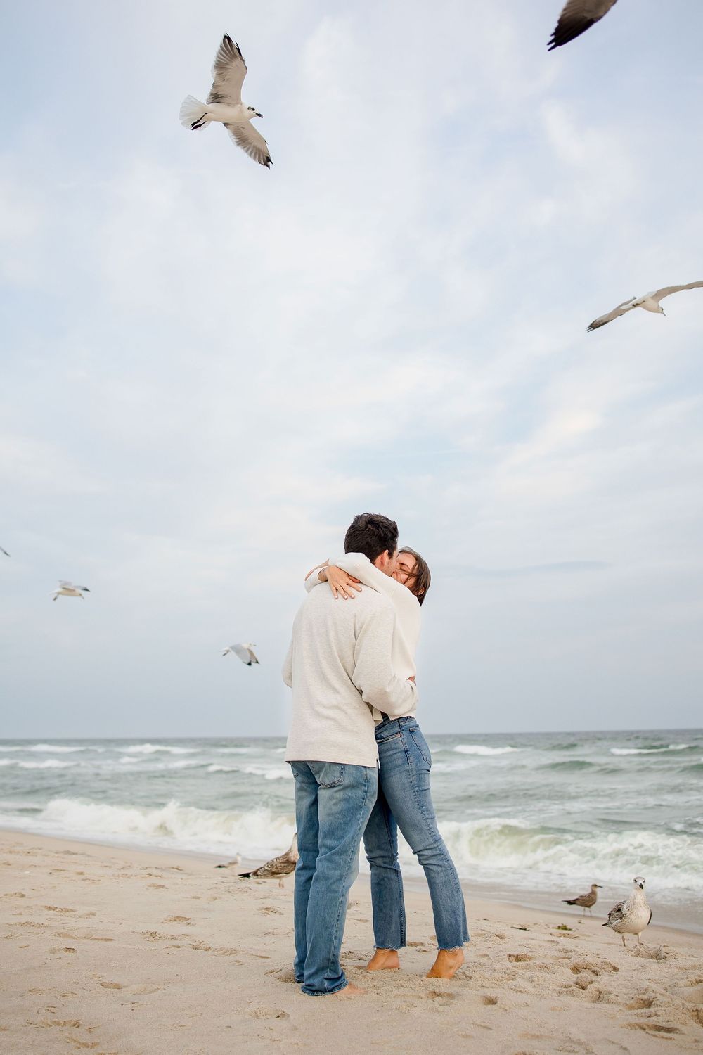 Two people in jeans stand on a beach watching seagulls fly over ocean waves on an overcast day.
