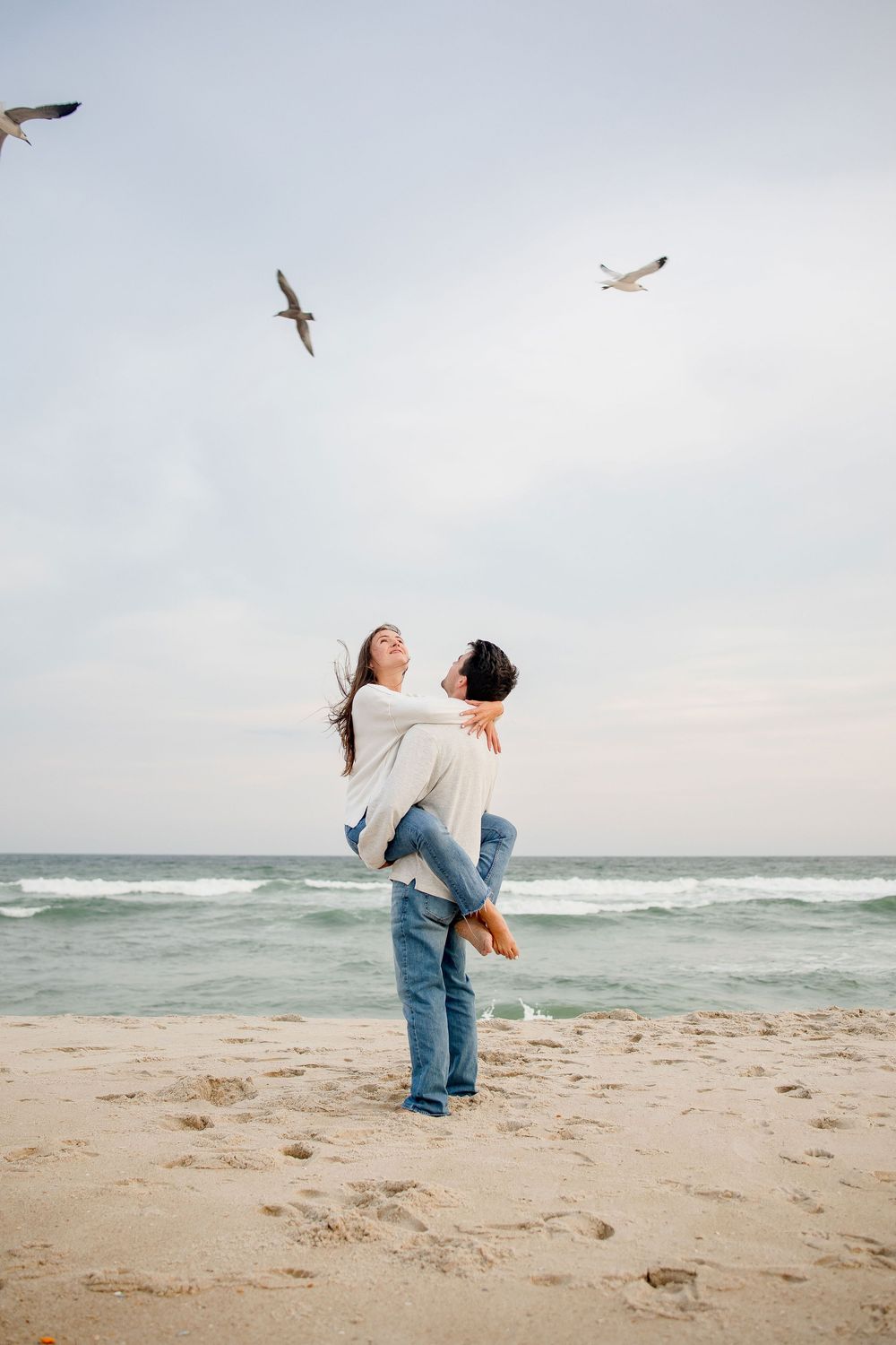 Two people share a romantic moment on a beach while seagulls fly overhead against a cloudy sky.