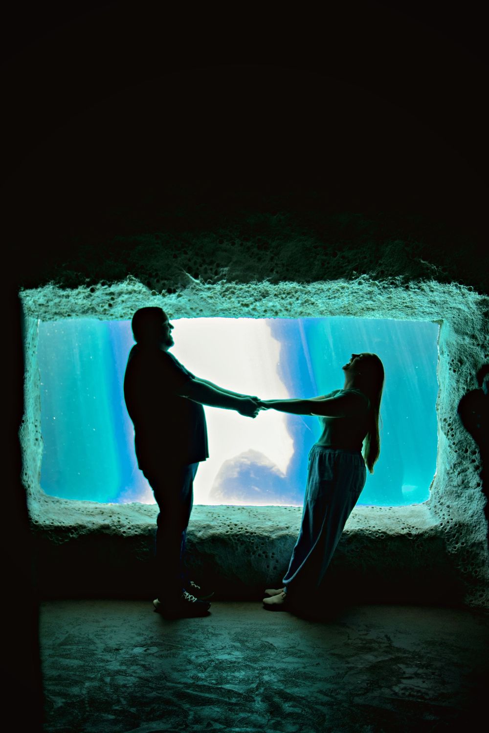 Silhouette of a couple holding hands in front of a glowing blue aquarium window in a dark room.