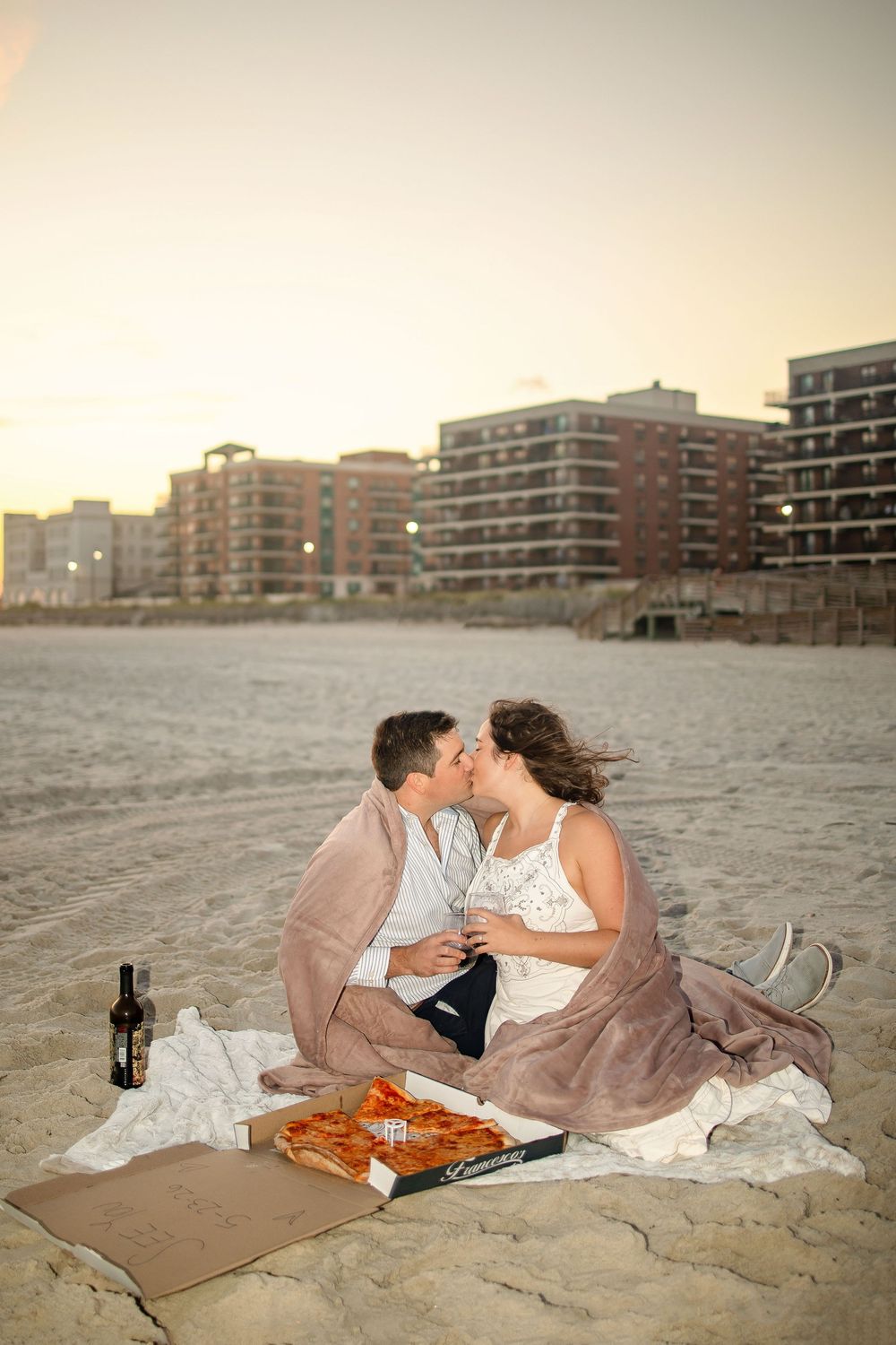 Romantic beach picnic at sunset with couple lounging on blankets near oceanfront condominiums.