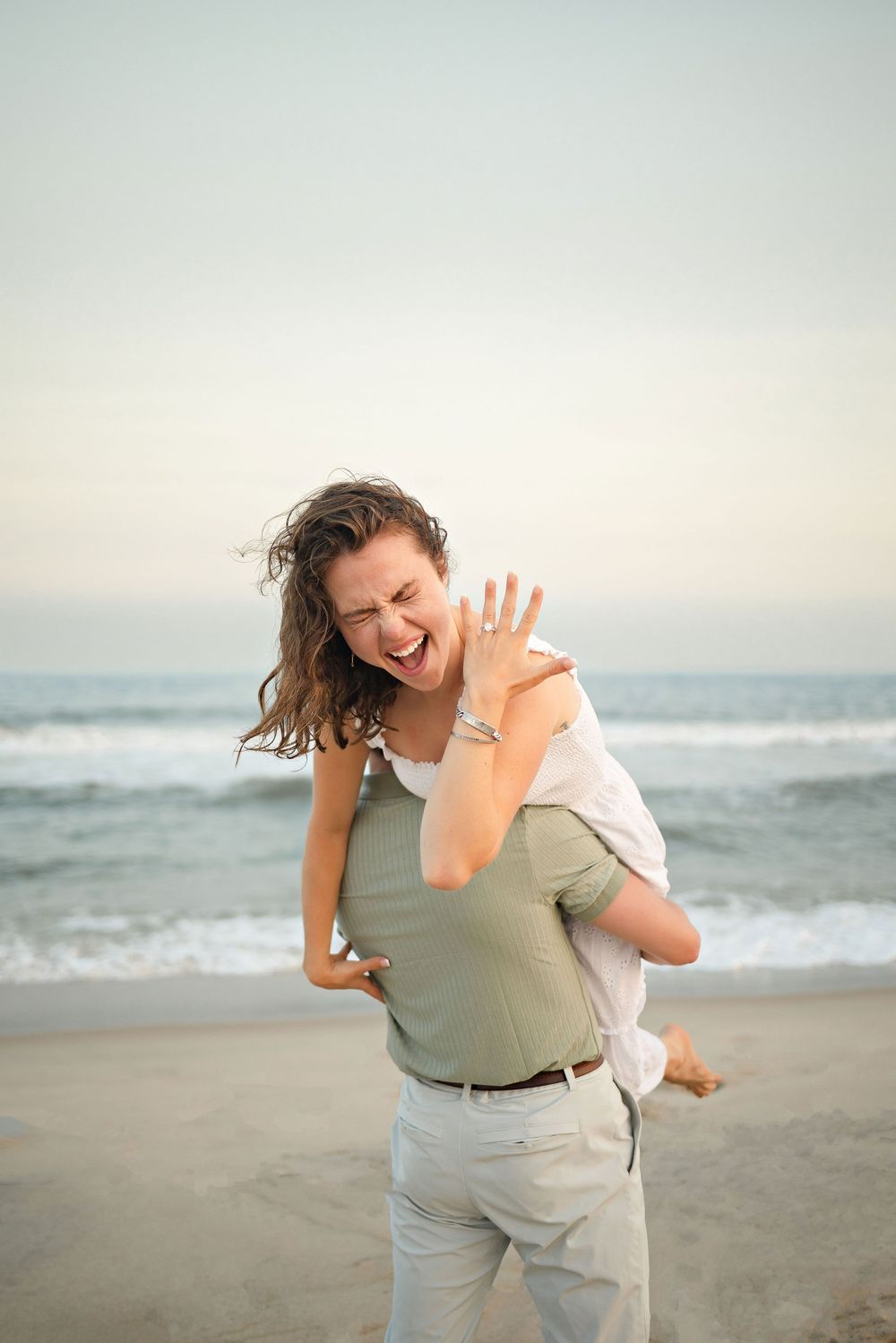 Joyful woman showing engagement ring while being carried by partner on a beach at sunset.
