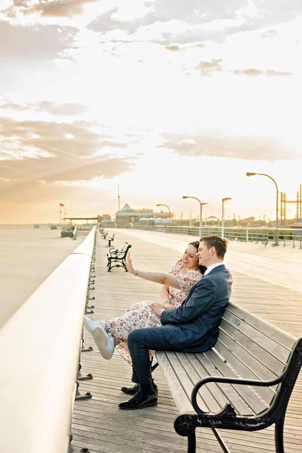 Couple sitting on boardwalk bench at sunset, taking a selfie, with amusement park lights glowing in the background.