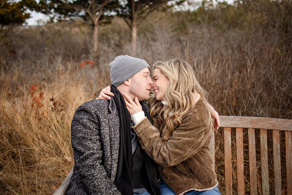 A romantic winter moment shared on a wooden bench surrounded by dried autumn grasses and bare trees.