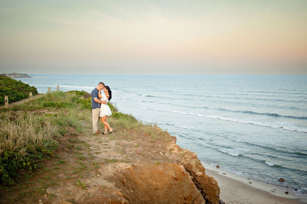 Couple embracing on a coastal cliff at sunset with ocean waves and sandy beach in the background.