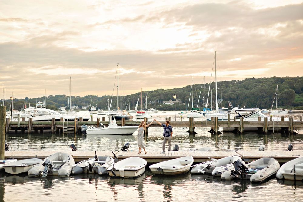 Two people stand on a dock at a marina with sailboats and motorboats at sunset.