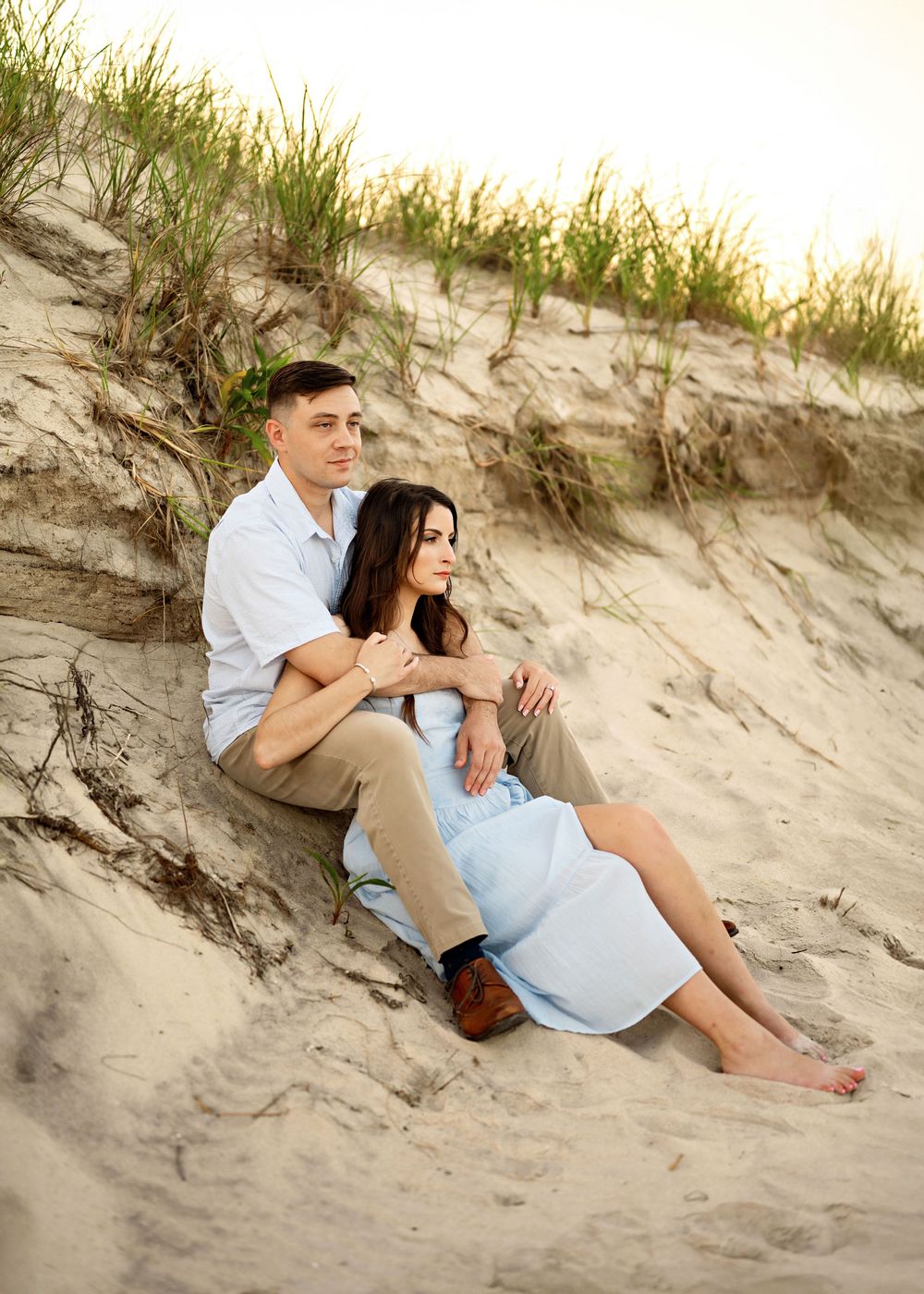 A couple sits intimately together on sandy beach dunes at sunset during a romantic portrait session.