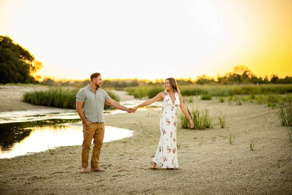 A couple holds hands on a sandy beach at sunset while wearing casual summer attire near a tidal pool.
