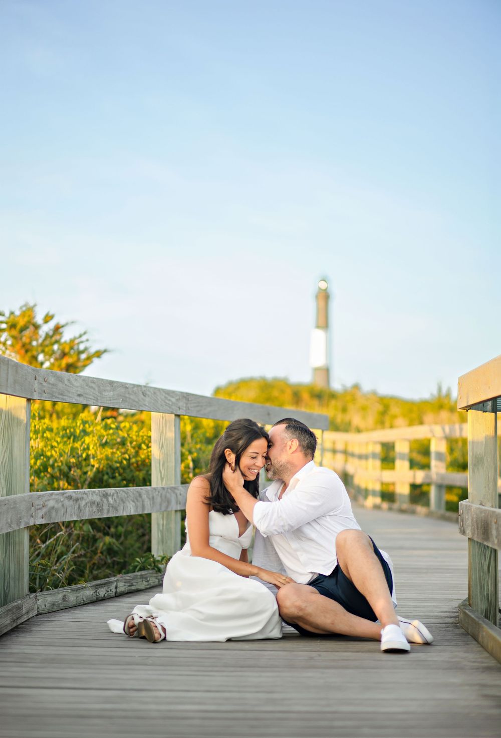 Couple sitting on a boardwalk near a lighthouse, sharing a romantic moment surrounded by golden wildflowers.