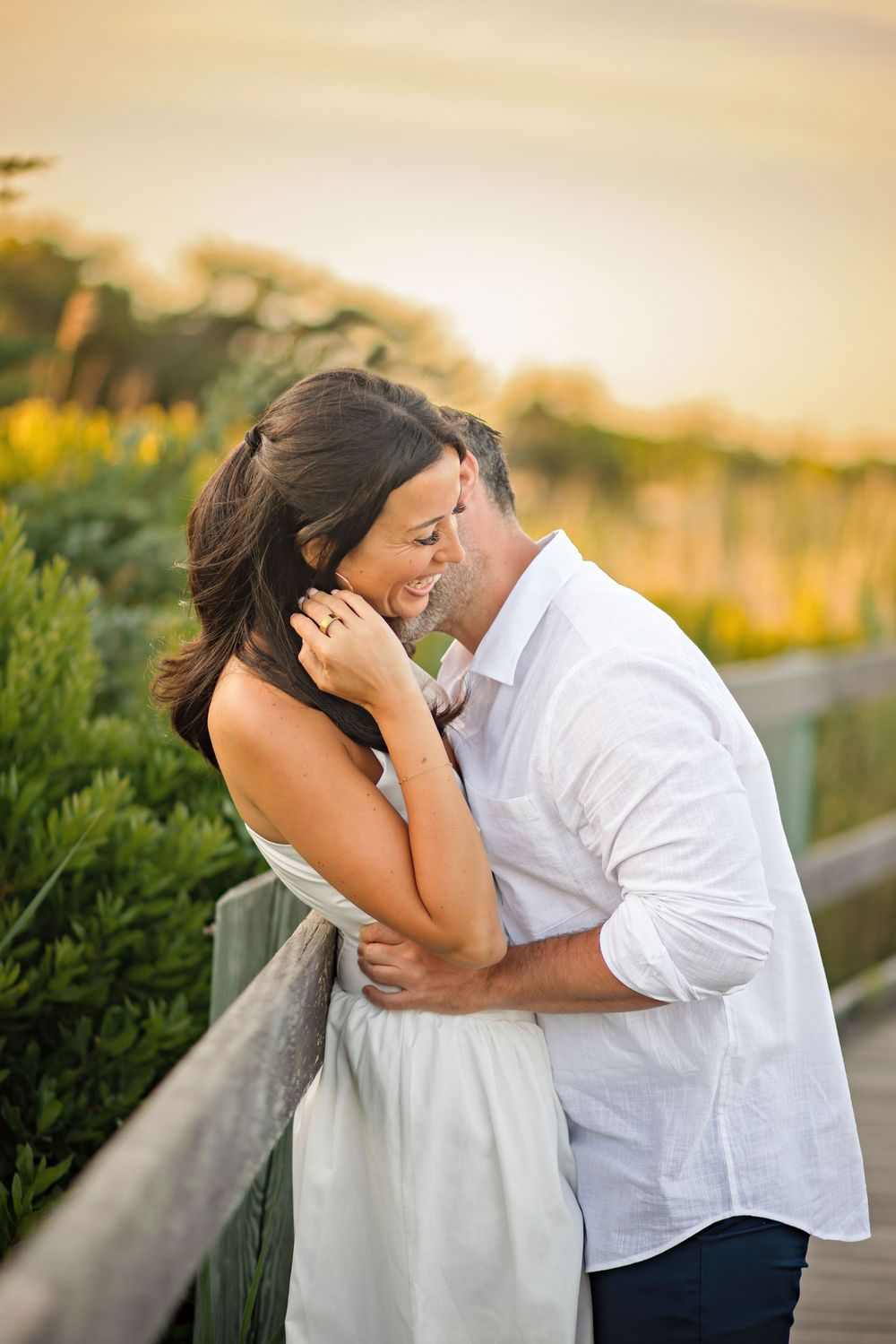 Couple sharing a romantic embrace on a wooden boardwalk at golden sunset, surrounded by lush greenery.