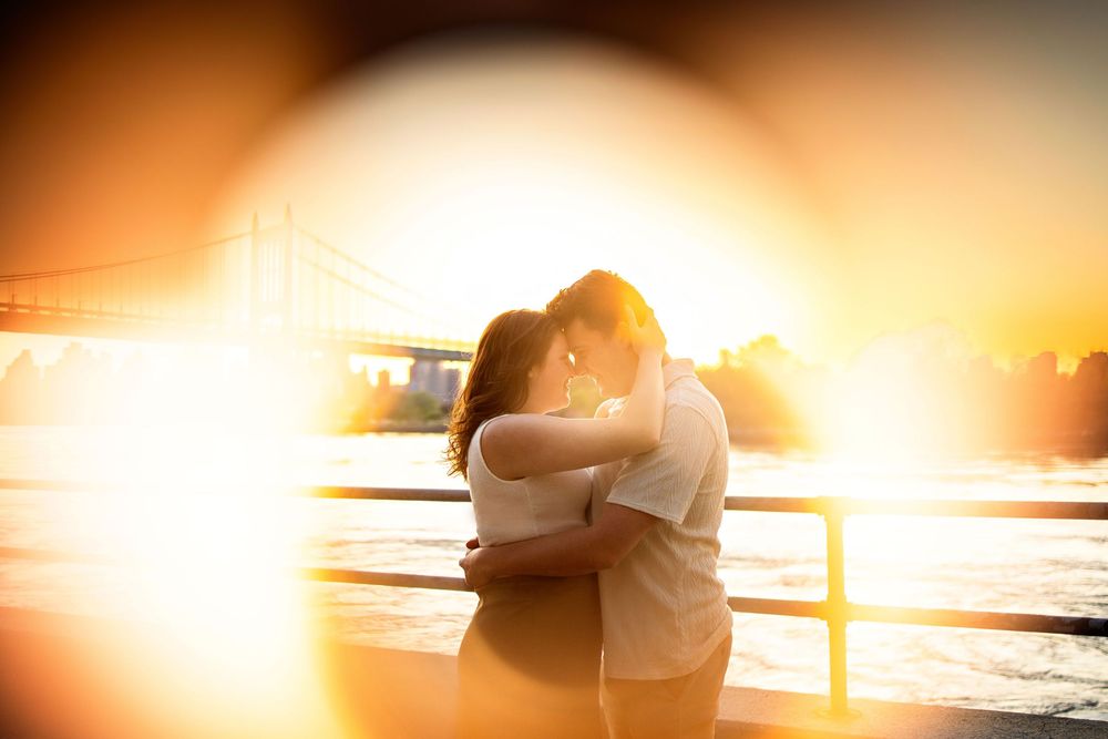 A couple embraces in a romantic silhouette against a golden sunset over water with lens flare effect.