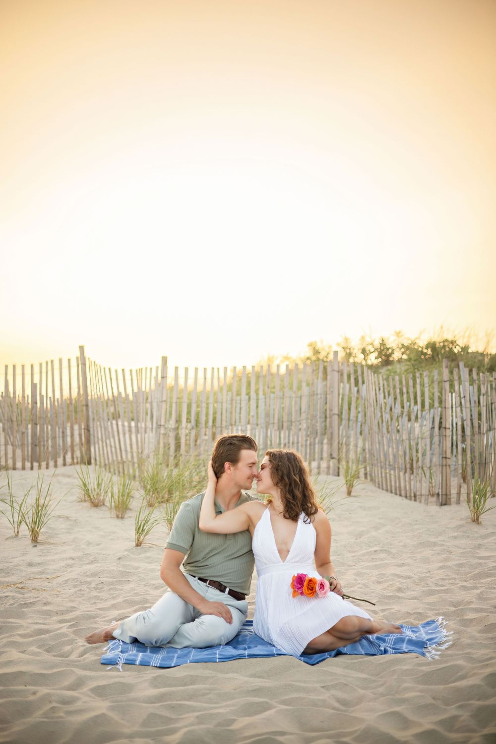 Couple sharing a romantic kiss on a blanket at a sandy beach during golden sunset near a wooden fence.