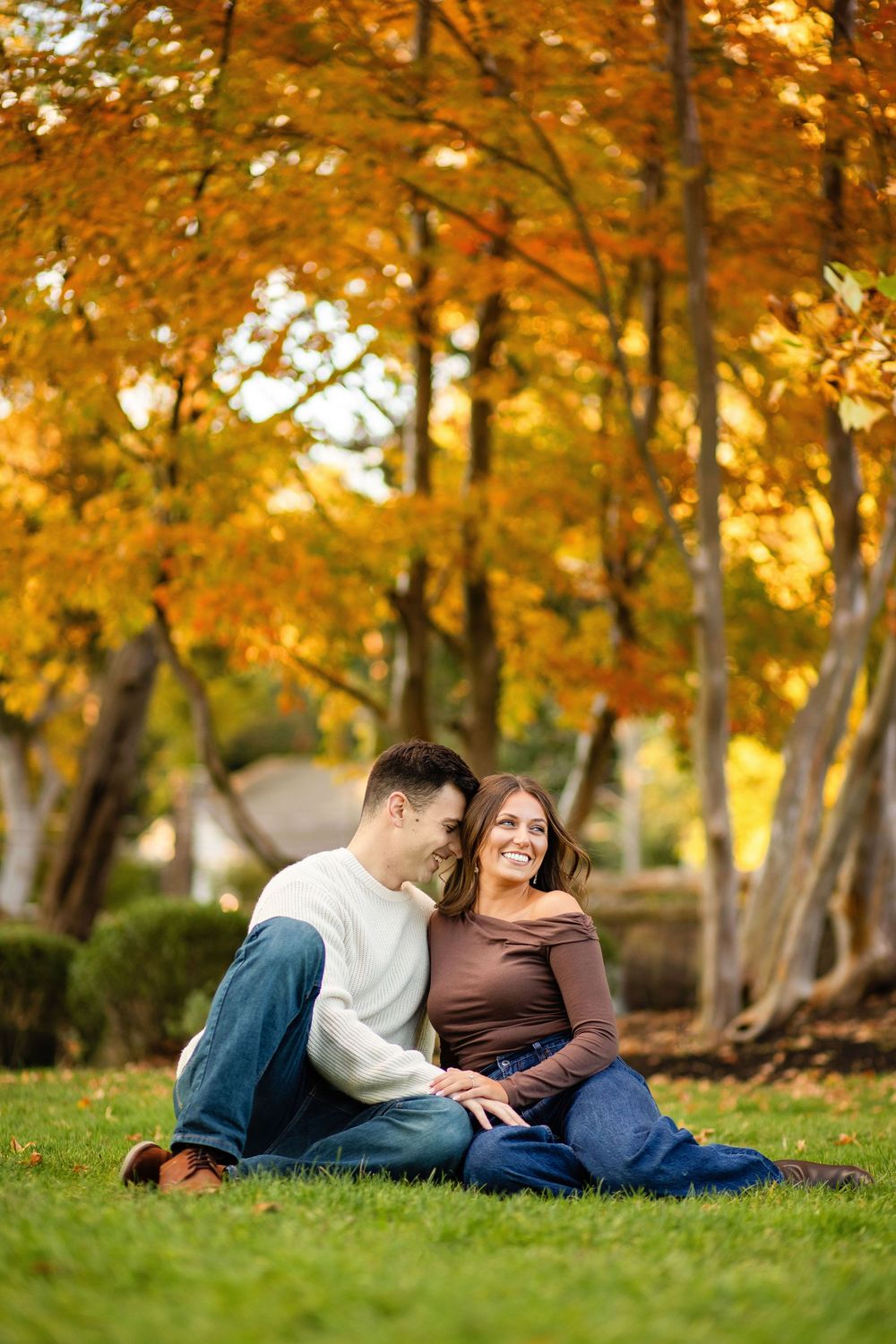 A couple shares an intimate moment while lying on grass surrounded by golden autumn trees at sunset.