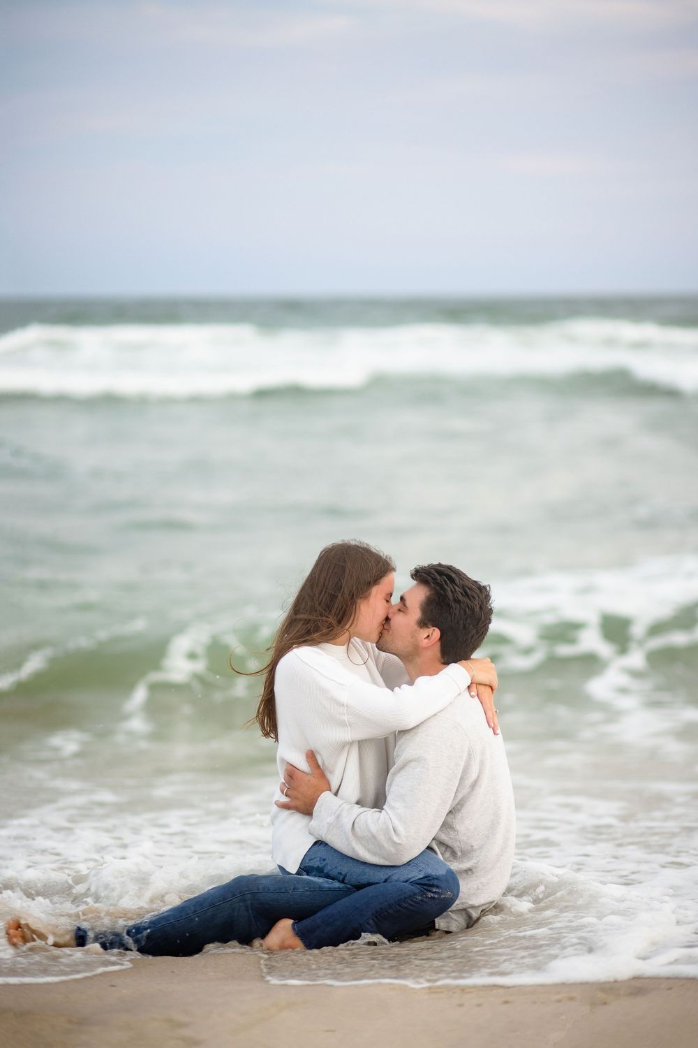 A couple shares a romantic moment sitting together on a sandy beach as ocean waves roll in behind them.
