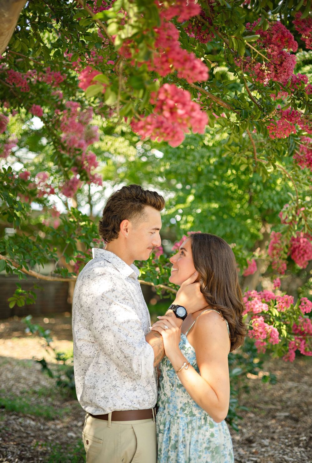 A romantic couple gazes at each other under blooming pink crape myrtle trees in a sun-drenched outdoor setting.