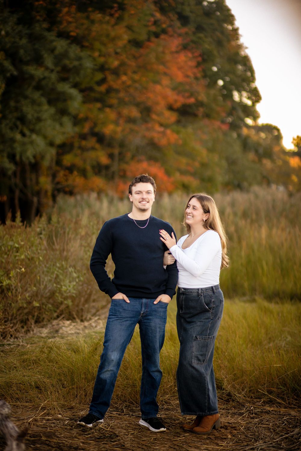 A couple standing together in a field with fall colors and trees in the background.