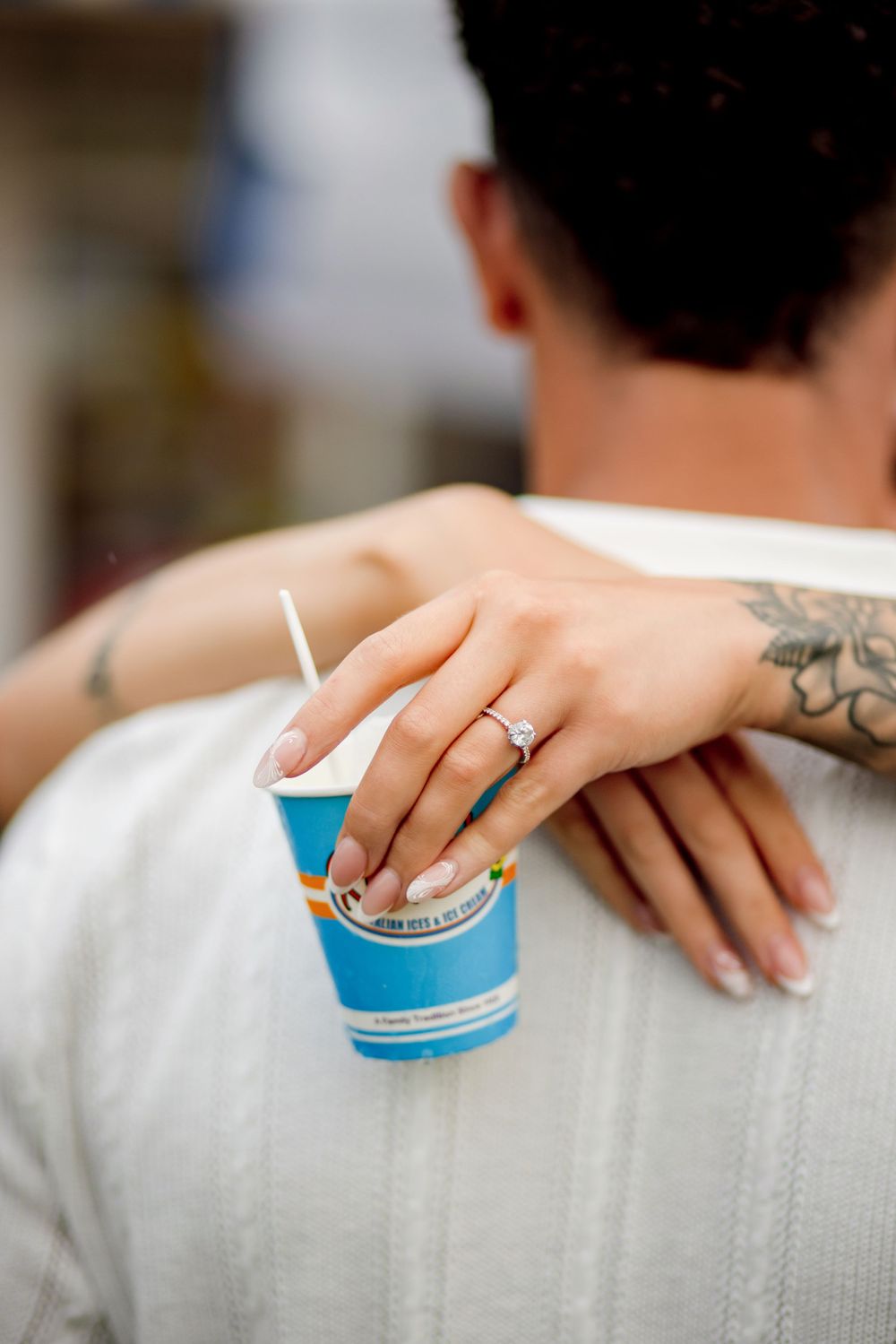 Close-up of tattooed hands holding an Italian ice cup with an engagement ring visible.