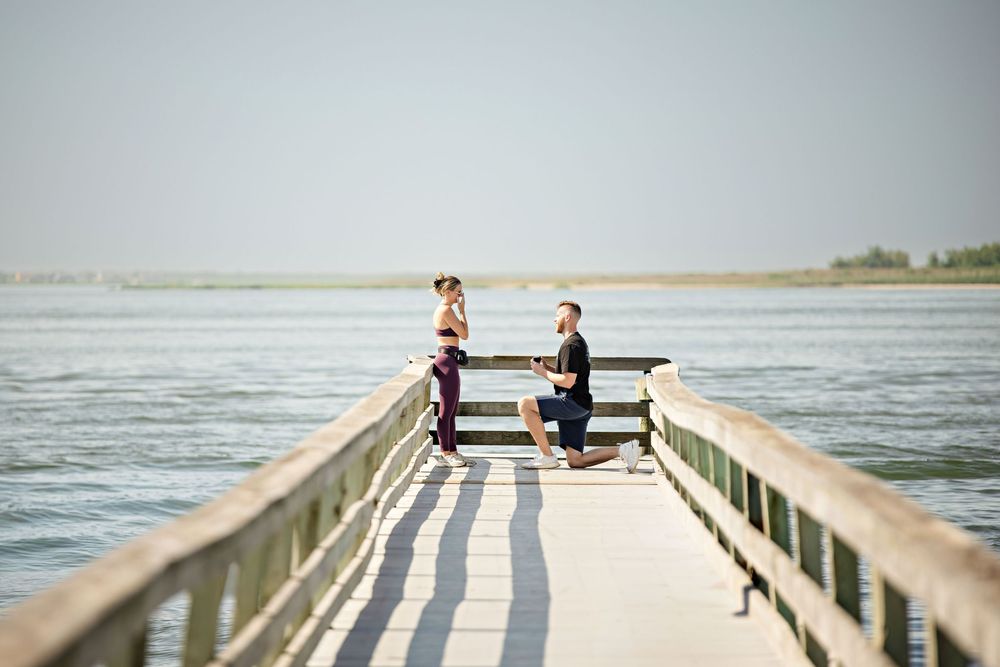 Man proposes on a wooden pier over calm water, woman reacts with surprise on a sunny day.